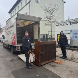 Dwóch mężczyzn w czarnych kurtkach i spodniach przenosi zabezpieczone pianino z ciężarówki z logo firmy przeprowadzkowej na chodnik. W tle budynek i ogrodzenie z banerem reklamowym.
