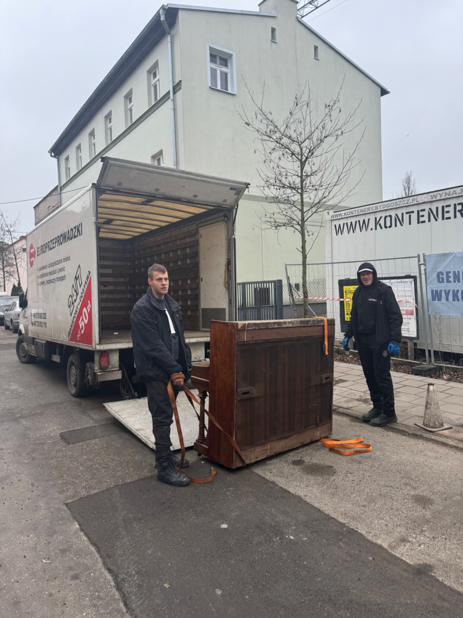 Dwóch mężczyzn w czarnych kurtkach i spodniach przenosi zabezpieczone pianino z ciężarówki z logo firmy przeprowadzkowej na chodnik. W tle budynek i ogrodzenie z banerem reklamowym.