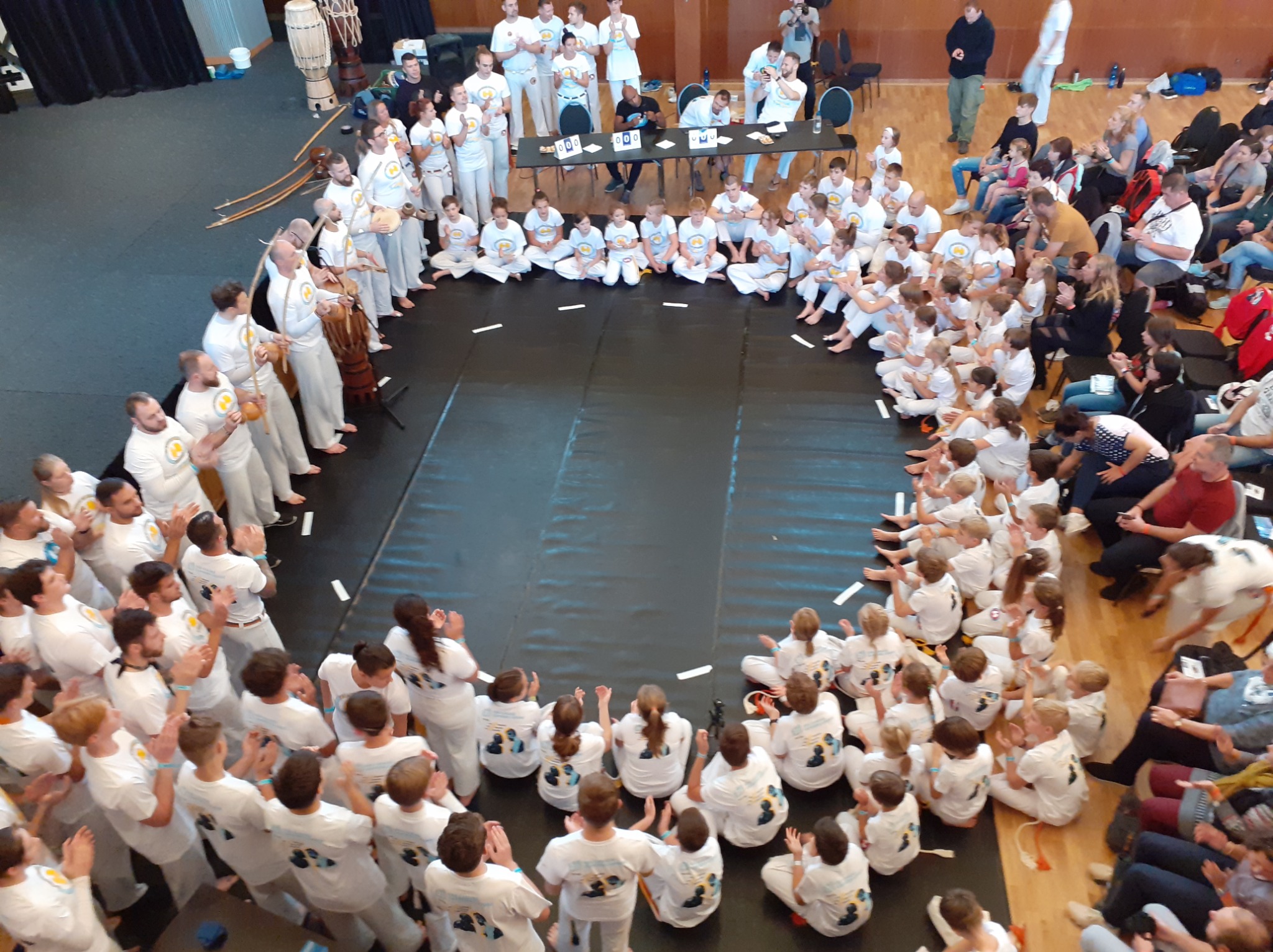 Duża grupa osób w białych strojach, część siedzi w kręgu na podłodze, część stoi z instrumentami, obserwatorzy na krzesłach i podłodze, widok z góry, prawdopodobnie występ capoeira.