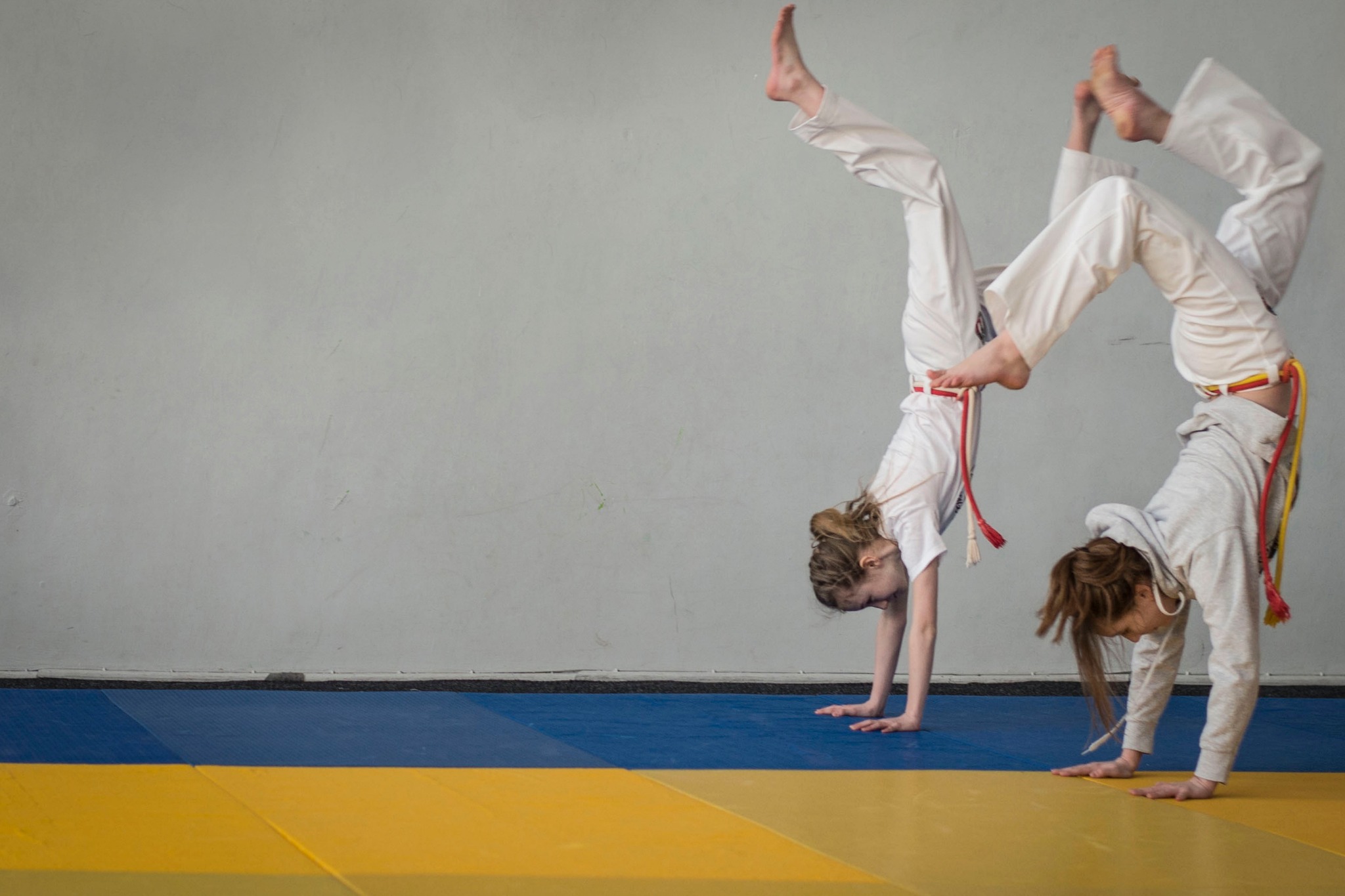 Dwie młode kobiety w białych strojach ćwiczą stójkę na rękach na niebiesko-żółtej macie w sali gimnastycznej, z widocznymi kolorowymi pasami. Jedna w bluzie z kapturem, druga w koszulce.