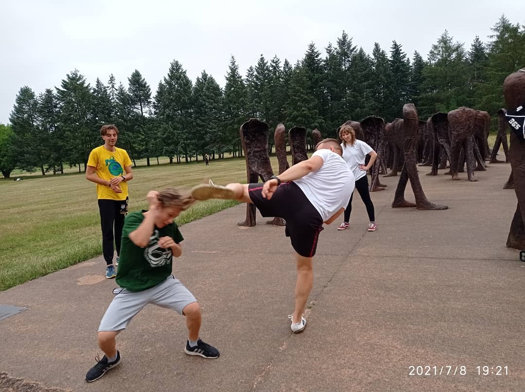 Mężczyzna w białej koszulce wykonuje kopnięcie w głowę drugiego mężczyzny w zielonej koszulce na tle rzeźb plenerowych i drzew.