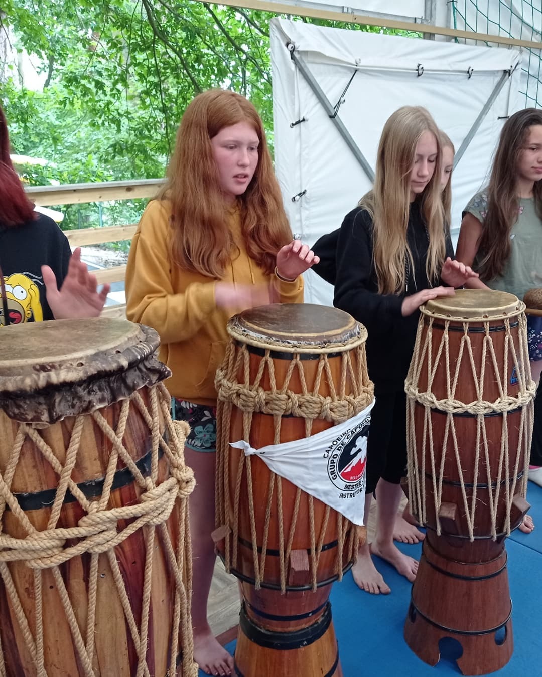 Grupa młodych osób w Poznaniu uczy się grać na berimbau podczas zajęć capoeira, widoczny biały proporczyk z logo grupy.