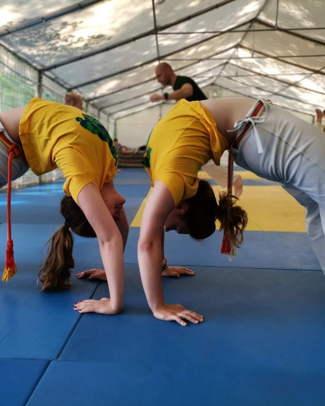 Dwie osoby w żółtych koszulkach wykonują mostek na niebiesko-żółtej macie, w tle widoczny instruktor i konstrukcja namiotu.