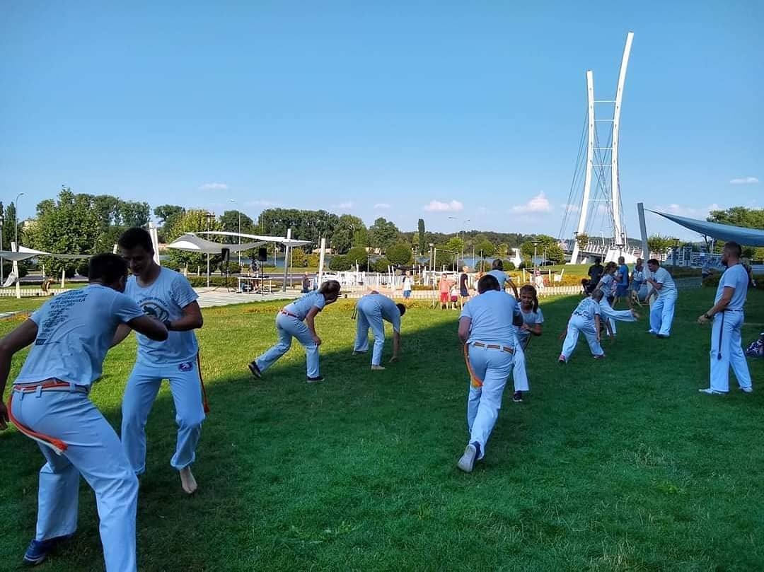 Grupa osób w białych strojach ćwiczy capoeirę na trawie w parku, z mostem w tle.