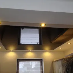 Nowoczesny sufit z czarną, błyszczącą wstawką i punktowym oświetleniem LED, widoczny fragment pomieszczenia w trakcie remontu z zakrytymi meblami.