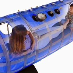 Kobieta i dziecko wewnątrz przenośnej, niebieskiej komory hiperbarycznej z manometrem i czarnymi podkładkami pod spodem.
