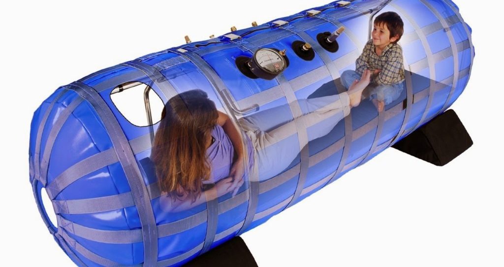 Kobieta i dziecko wewnątrz przenośnej, niebieskiej komory hiperbarycznej z manometrem i czarnymi podkładkami pod spodem.