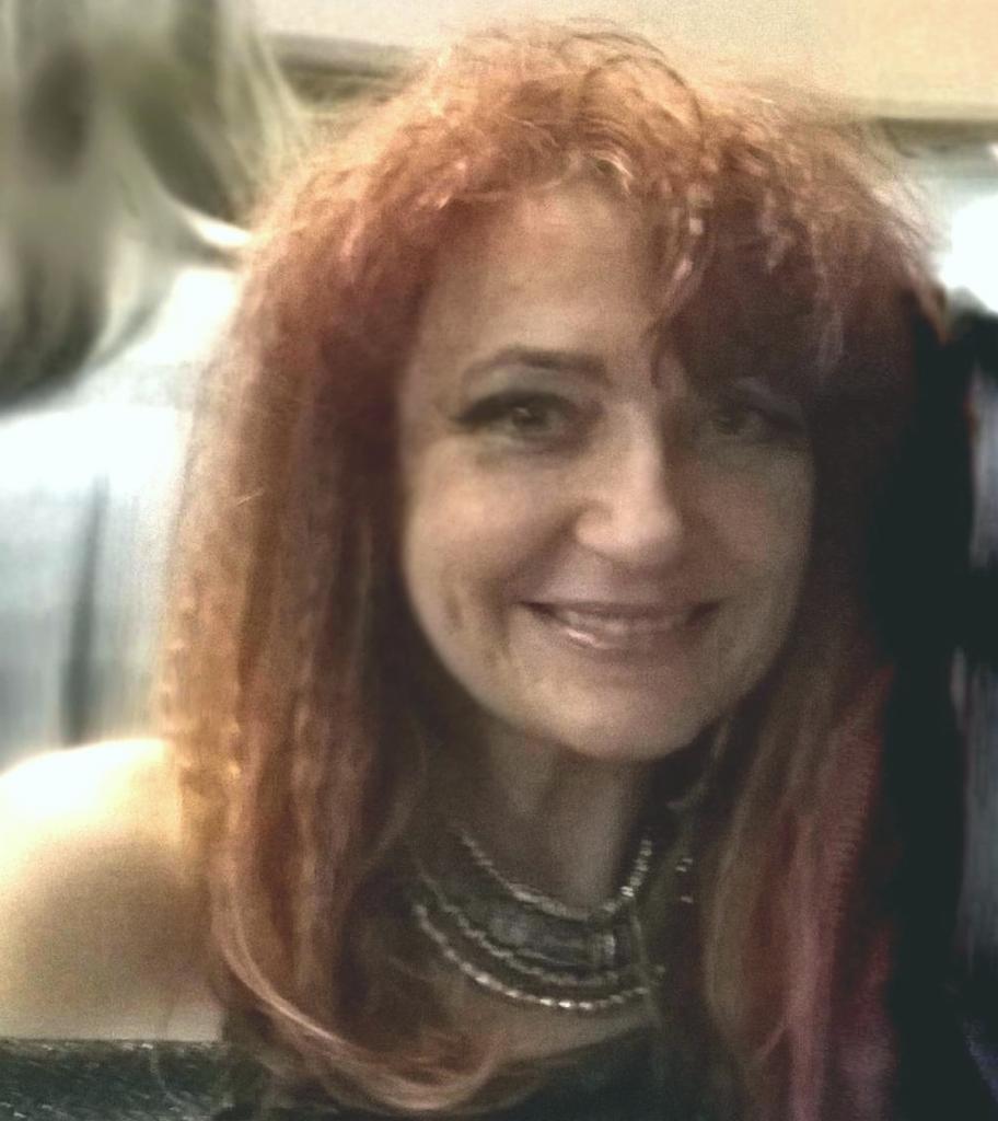 Portret uśmiechniętej kobiety z rudymi kręconymi włosami i naszyjnikiem. Ujęcie z bliska, miękkie oświetlenie, rozmyte tło. Delikatny makijaż.