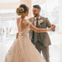 Para młoda tańcząca w jasnym wnętrzu sali balowej, panna młoda w koronkowej sukni z odkrytymi plecami, pan młody w szarym garniturze w kratę i bordowym krawacie.