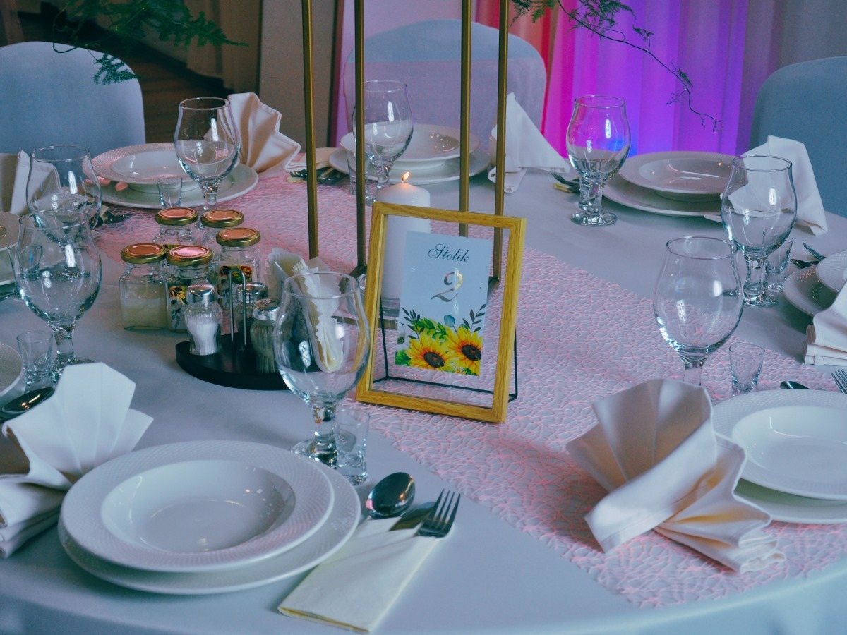 Elegancko nakryty stół z białymi talerzami, serwetkami, szklankami i dekoracjami, w tym słoiczkami z zawartością, solniczką i pieprzniczką oraz ramką ze słonecznikami i numerem stołu