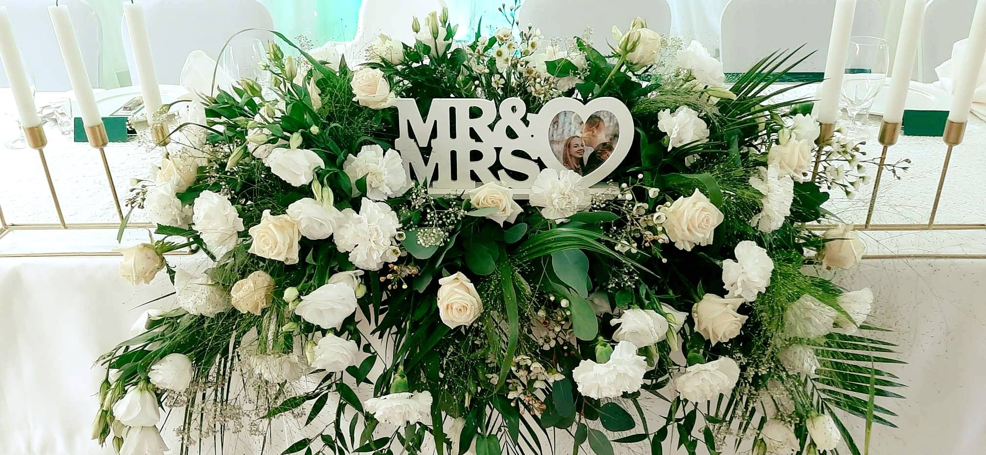 Dekoracja stołu weselnego z białych róż, goździków i zielonych liści, z napisem 'Mr & Mrs' i ramką w kształcie serca ze zdjęciem pary młodej.