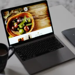 Laptop wyświetla stronę internetową z wizerunkiem oliwek w drewnianej misce, obok kubek z kawą, notatnik i telefon na białym biurku.