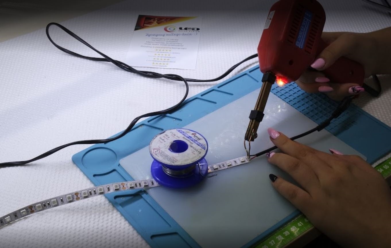 Podłączenie przewodów do taśmy LED za pomocą lutownicy pistoletowej; widoczna cyna, mata robocza i ulotka reklamowa.