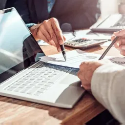 Spotkanie biznesowe przy biurku z laptopem, kalkulatorem i dokumentami; dłonie wskazują na wykresy, oświetlenie słoneczne