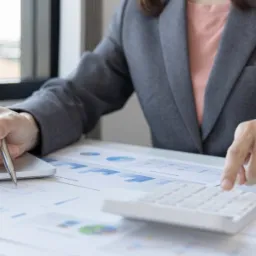 Kobieta w szarym żakiecie analizuje wykresy, używając kalkulatora i długopisu przy laptopie na biurku w jasnym pomieszczeniu.