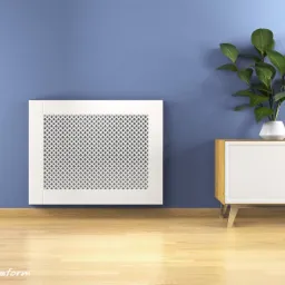 Biała, ażurowa osłona grzejnika z motywem rombów, zamontowana na niebieskiej ścianie, obok komody z rośliną doniczkową w nowoczesnym wnętrzu z drewnianą podłogą.