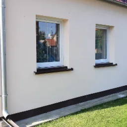 Jasna elewacja domu jednorodzinnego z dwoma oknami, brązowymi parapetami i listwą przypodłogową, widoczny fragment dachu z czerwoną dachówką, rynna i trawnik.