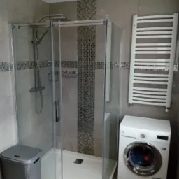 Nowoczesna łazienka z kabiną prysznicową, grzejnikiem drabinkowym i pralką, wykończona szarymi płytkami z dekoracyjnym pasem mozaiki.