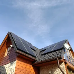 Panele słoneczne na dachu domu z drewnianą elewacją i kamiennym wykończeniem, antena satelitarna, błękitne niebo z chmurami w tle.
