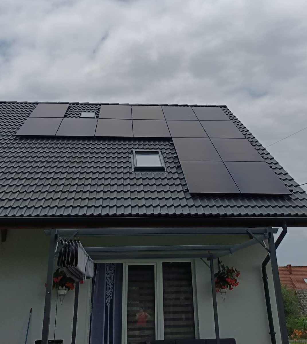 Czarny dach z dachówki ceramicznej z zamontowanymi panelami słonecznymi i oknami dachowymi, widok z dołu na szarym, pochmurnym tle.