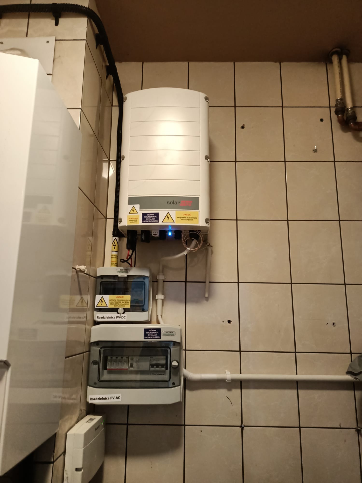 Zamontowana rozdzielnica PV-DC i PV-AC oraz inwerter fotowoltaiczny SolarEdge na ścianie z płytek, widoczne przewody elektryczne i oznaczenia ostrzegawcze.