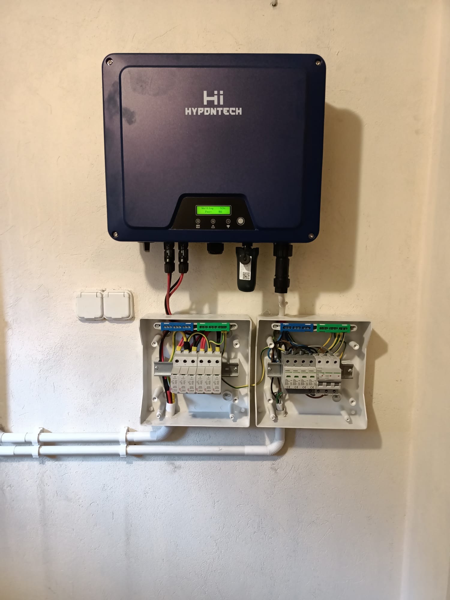 Niebieski falownik Hypontech zamontowany nad dwoma otwartymi skrzynkami elektrycznymi z bezpiecznikami i okablowaniem, na tle kremowej ściany z widocznymi rurami PCV.