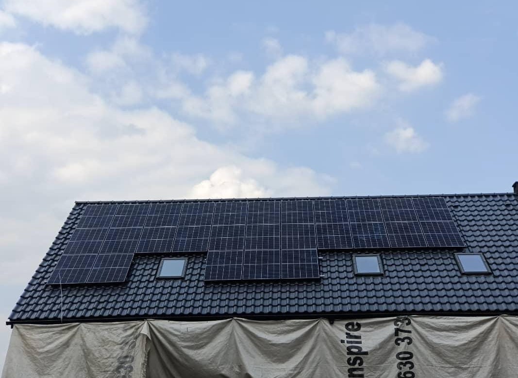 Panele słoneczne na dachu domu z dachówką ceramiczną, widoczne okna dachowe i zabezpieczenie folią na dole dachu, błękitne niebo z chmurami w tle.