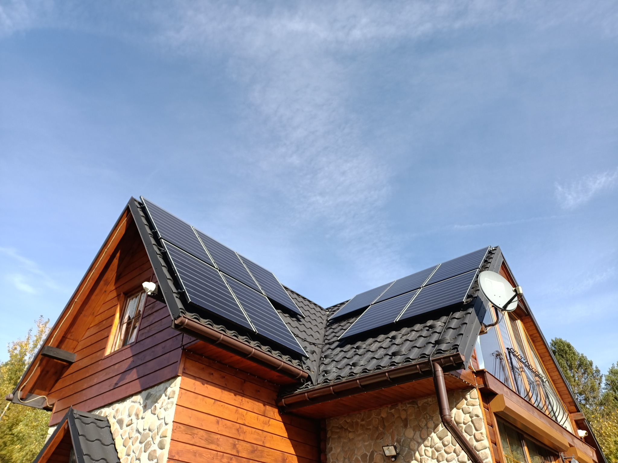 Panele słoneczne na dachu domu z drewnianą elewacją i kamiennym wykończeniem, antena satelitarna, błękitne niebo z chmurami w tle.