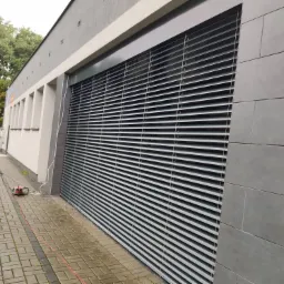 Szare poziome żaluzje zewnętrzne zamontowane na ścianie budynku z szarych płyt, widoczny pracownik z narzędziem na chodniku z kostki.