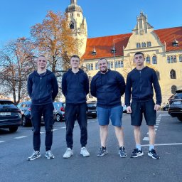 Helper transport & przeprowadzki - Czterech mężczyzn w ciemnych bluzach stoi na parkingu przed budynkiem z czerwoną dachówką i wieżą. W tle zaparkowane samochody i błękitne niebo.