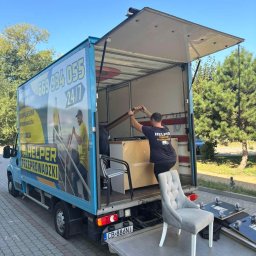 Helper transport & przeprowadzki - Pracownik w trakcie załadunku mebli do busa z rampą, na której stoi krzesło. Na boku pojazdu reklama firmy przeprowadzkowej z numerem telefonu.