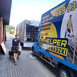 Helper transport & przeprowadzki - Pracownik firmy przeprowadzkowej Helper transportuje wózek z zapakowanymi przedmiotami do otwartej ciężarówki. W tle niebieski bus z reklamą firmy.