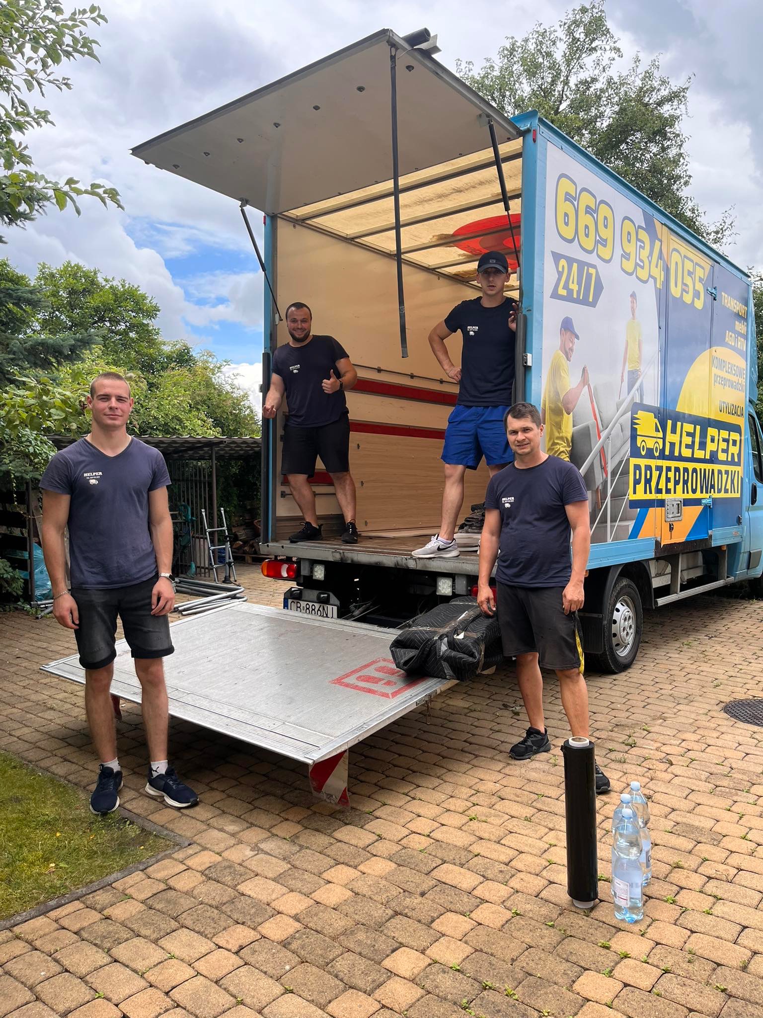 Ekipa przeprowadzkowa Helper pozuje na tle otwartego busa z reklamą firmy. Widoczny numer rejestracyjny pojazdu i butelki wody. Częściowo widoczne drzewa w tle.