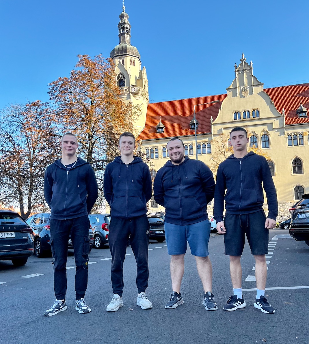 Czterech mężczyzn w ciemnych bluzach pozuje na tle budynku z wieżą i aut. Jasne niebo, jesienne drzewa. Ujęcie z dołu, podkreślające wysokość budowli.