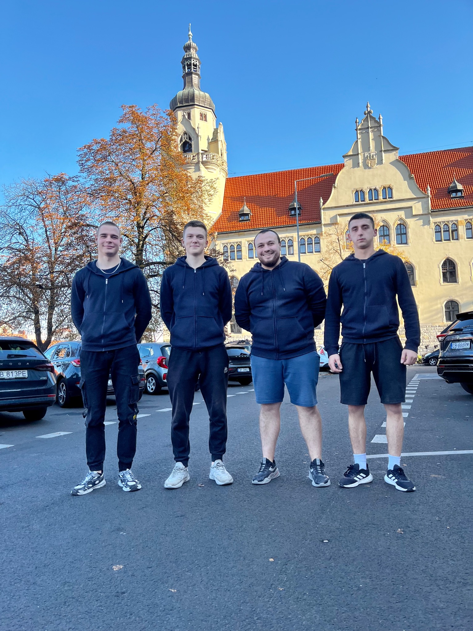 Czterech mężczyzn w ciemnych bluzach stoi na parkingu przed budynkiem z czerwoną dachówką i wieżą. W tle zaparkowane samochody i błękitne niebo.