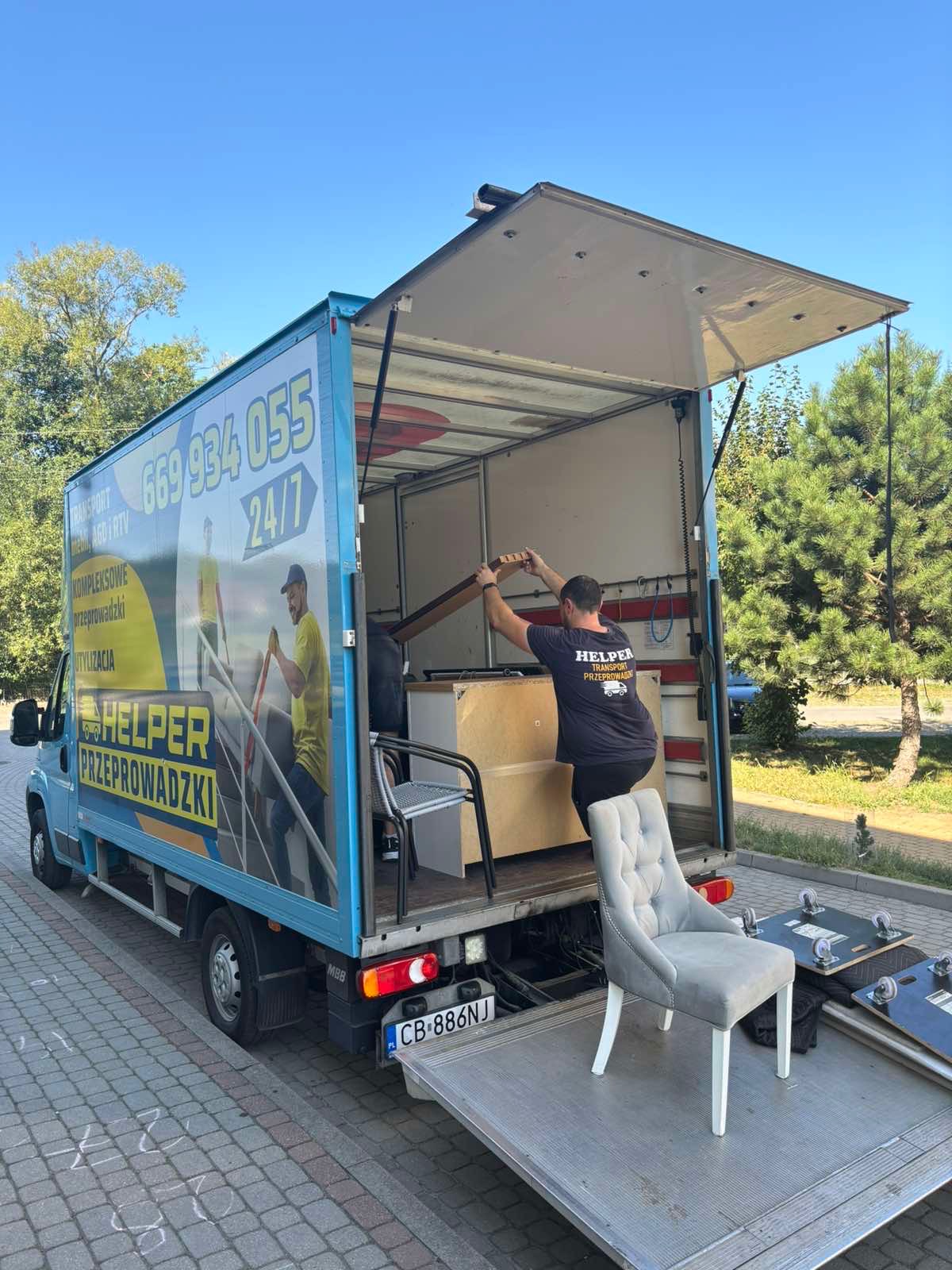 Pracownik w trakcie załadunku mebli do busa z rampą, na której stoi krzesło. Na boku pojazdu reklama firmy przeprowadzkowej z numerem telefonu.