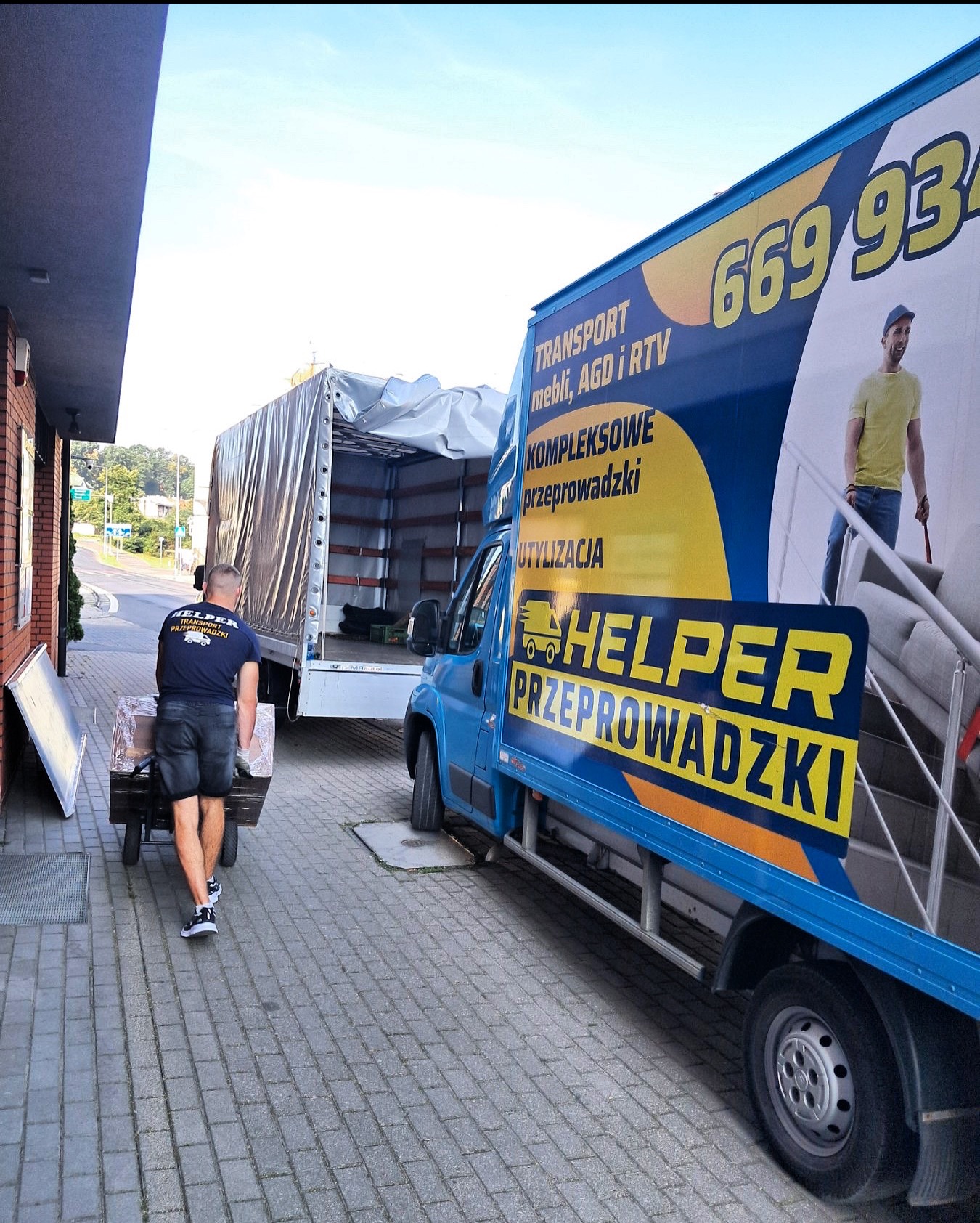 Pracownik firmy przeprowadzkowej Helper transportuje wózek z zapakowanymi przedmiotami do otwartej ciężarówki. W tle niebieski bus z reklamą firmy.