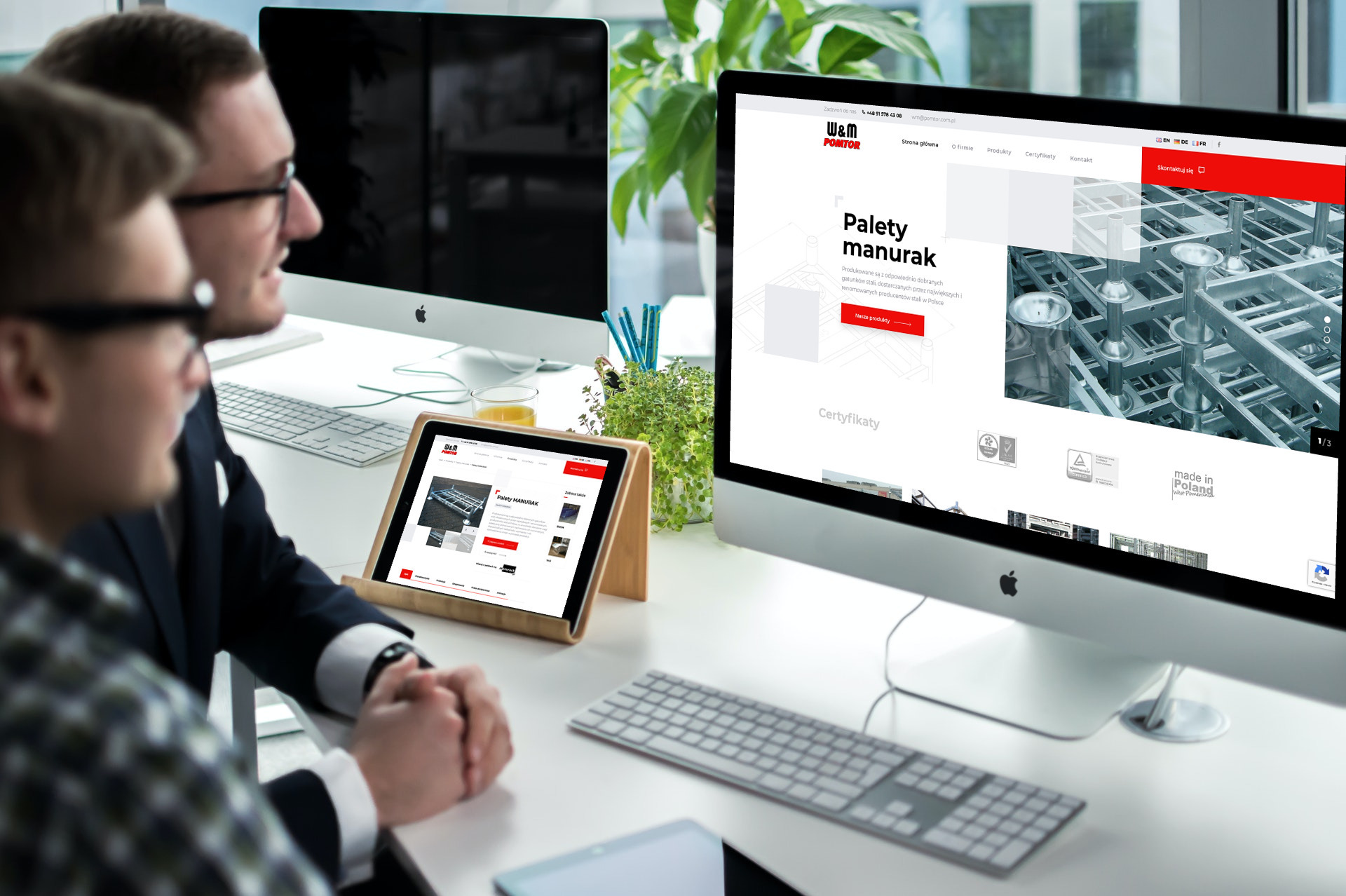 Dwóch mężczyzn w okularach patrzy na ekrany komputerów i tabletu prezentujące stronę internetową firmy W&M Pomtor, specjalizującej się w produkcji palet manurak. Na biurku rośliny doniczkowe...
