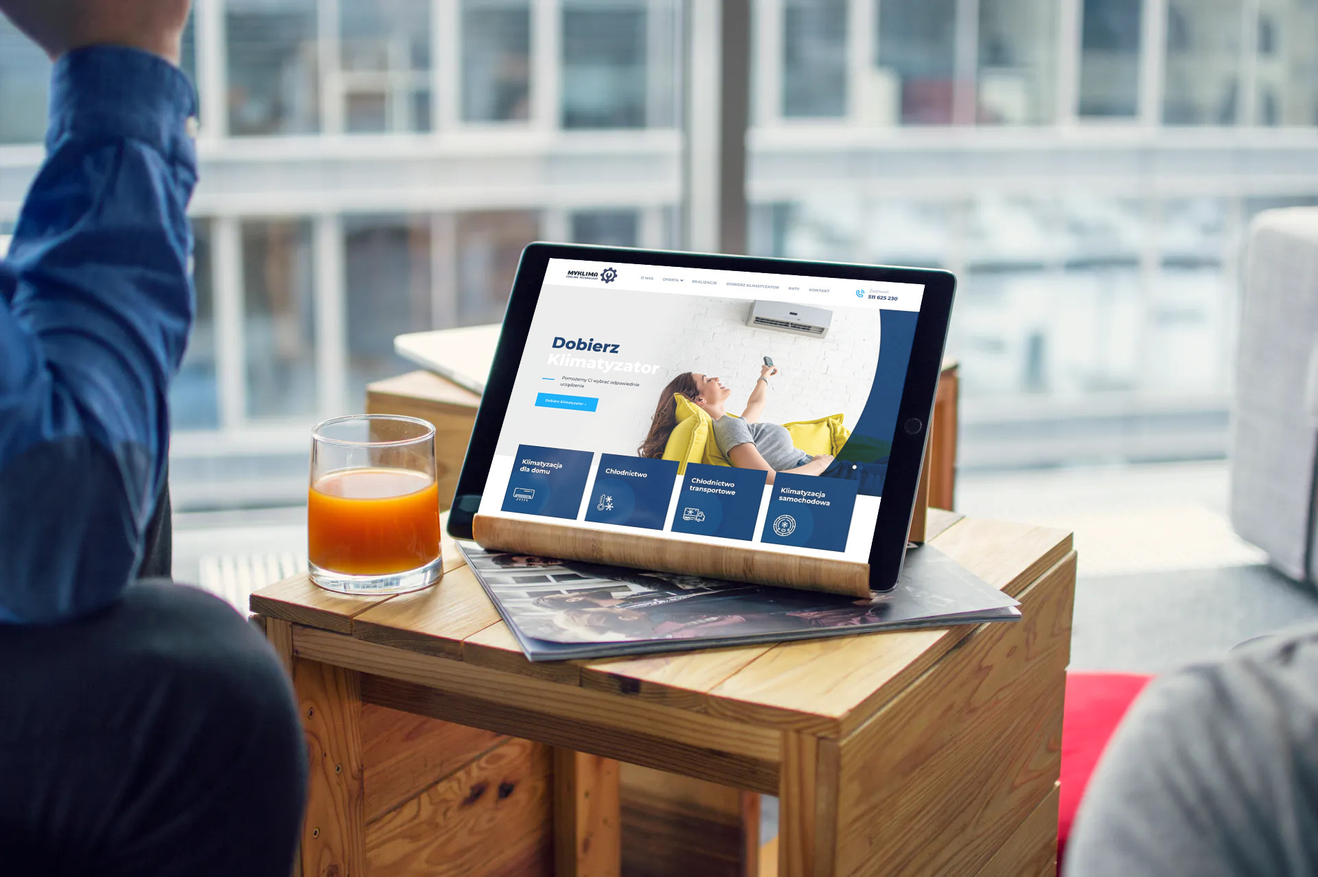 Tablet prezentuje responsywną stronę internetową firmy z branży klimatyzacji na drewnianym stoliku obok szklanki soku pomarańczowego, widok z perspektywy osoby siedzącej.
