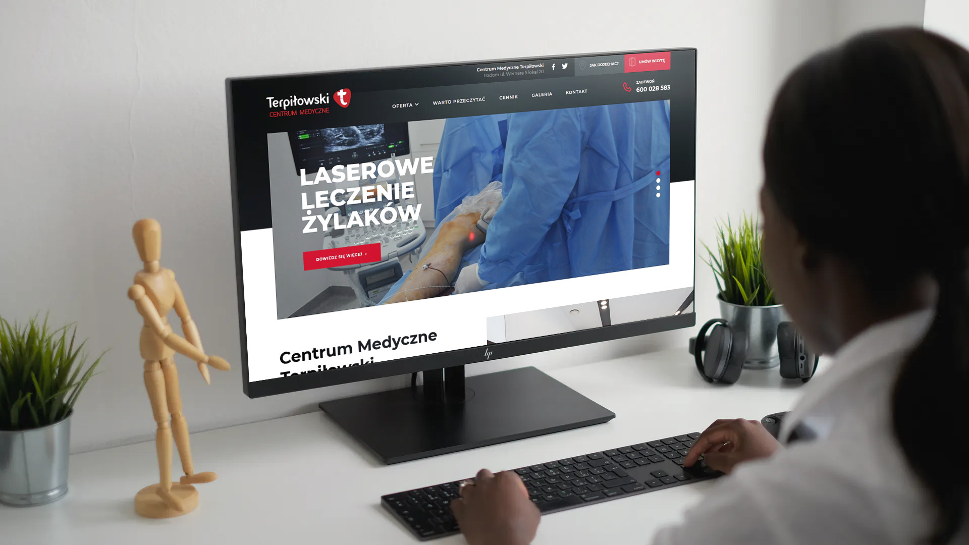 Kobieta w białym fartuchu przegląda na monitorze stronę internetową centrum medycznego Terpiłowski z ofertą laserowego leczenia żylaków. Na biurku stoi drewniany manekin, roślina w doniczce...