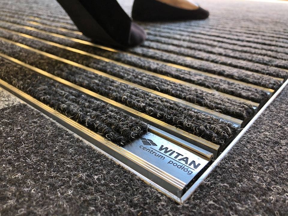 Szczegółowe ujęcie wycieraczki systemowej z logo Witan centrum podłóg, z widoczną nogą w czarnym balerinku, na tle szarej wykładziny dywanowej.