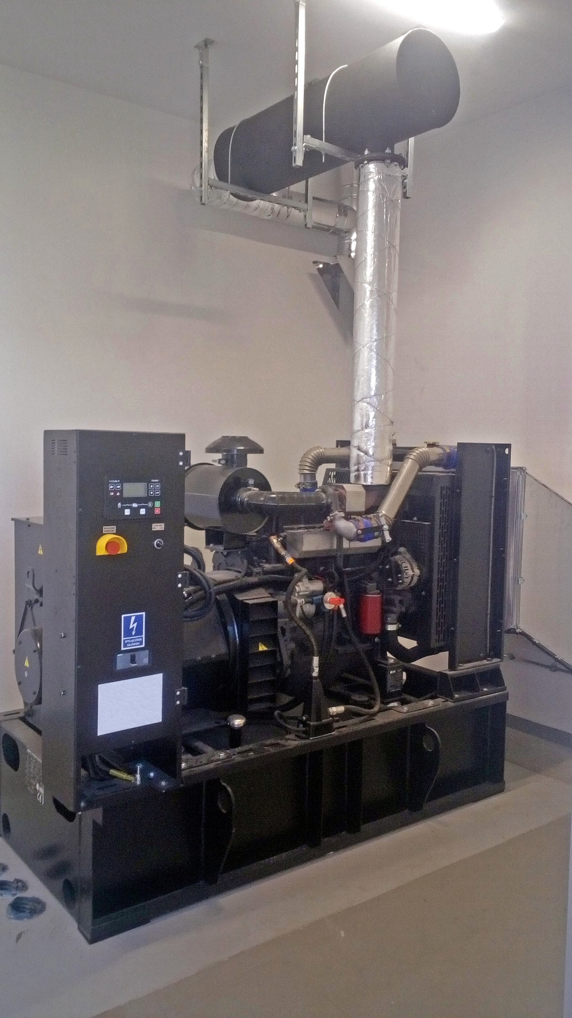 Stacjonarny agregat prądotwórczy z czarną obudową, panelem sterowania z wyświetlaczem i rurą wydechową owiniętą izolacją, zamontowany w pomieszczeniu z szarą podłogą i białą ścianą.