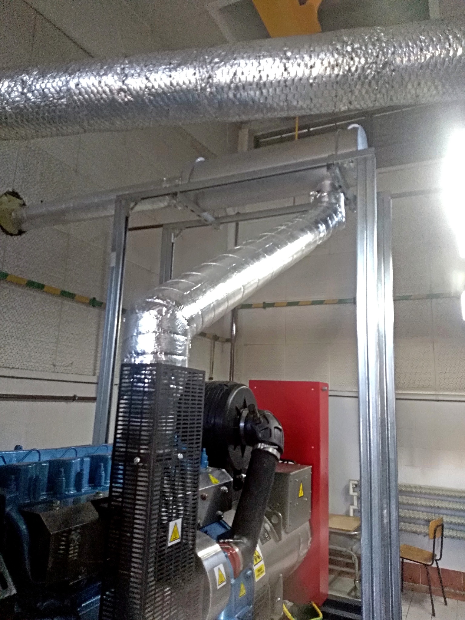 Wnętrze hali z widocznym systemem wentylacyjnym z izolowanymi rurami, generatorem awaryjnym z niebieskim silnikiem i czerwonym panelem sterowania oraz metalową konstrukcją nośną.