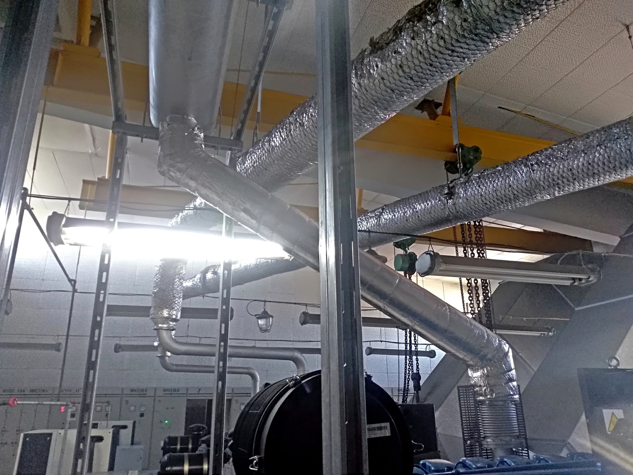 Wnętrze hali z widocznymi instalacjami wentylacyjnymi, rurami, podwieszonymi lampami, konstrukcją nośną sufitu i panelami sterowania urządzeniami.