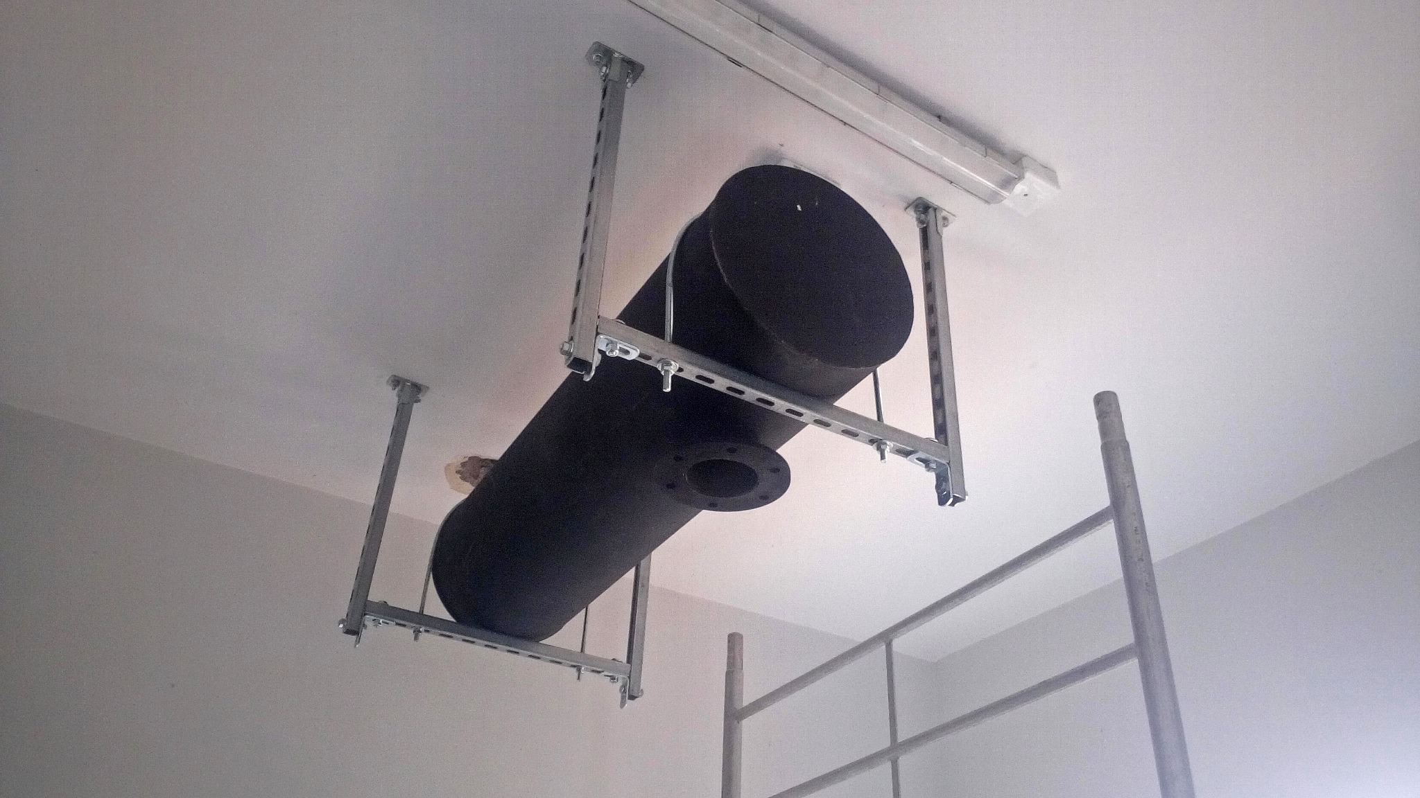 Czarny, cylindryczny zbiornik zamontowany pod sufitem na metalowej konstrukcji, widoczna flansza, drabina w tle.