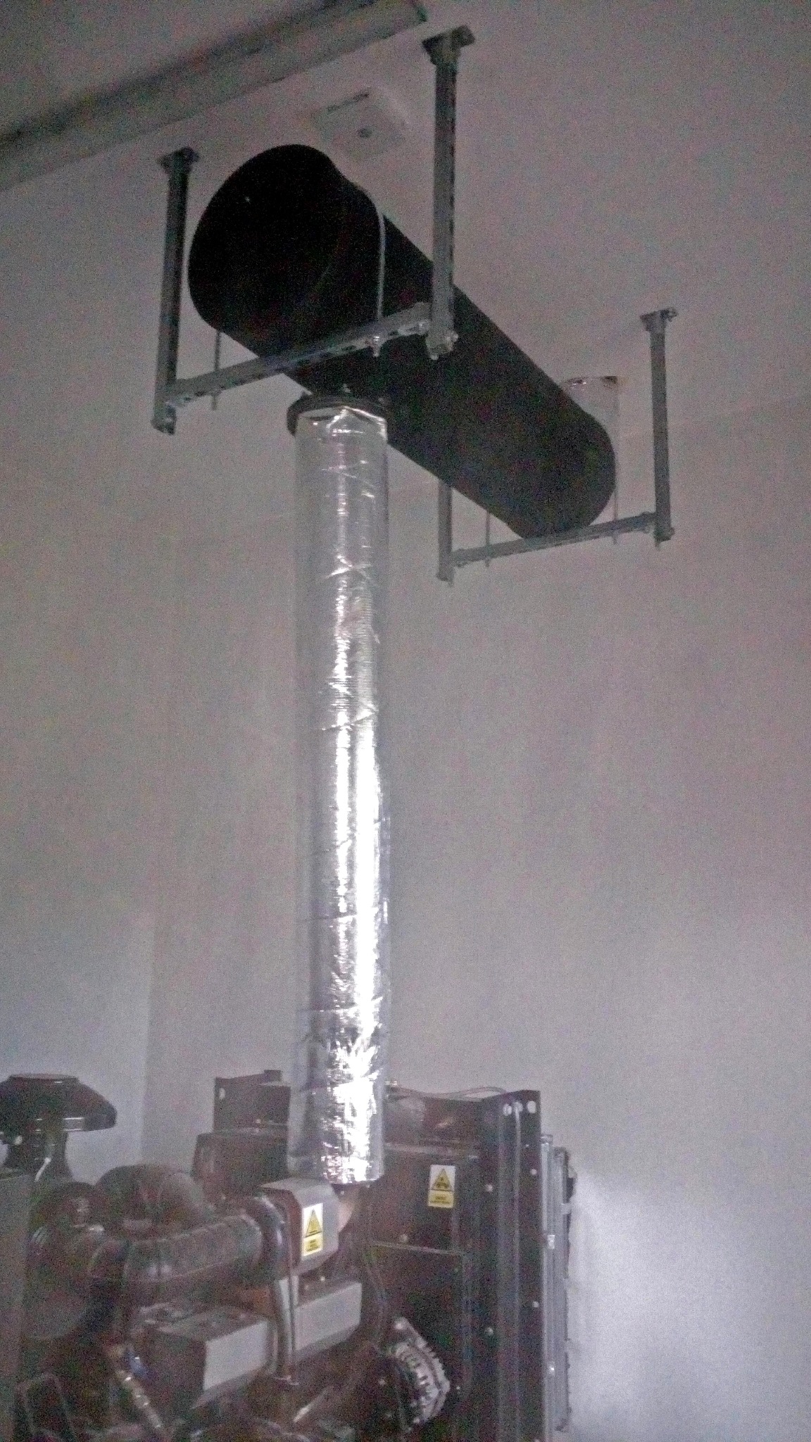 Czarny tłumik spalin agregatu przymocowany do sufitu za pomocą stalowej konstrukcji, z widoczną izolacją termiczną na rurze spalinowej.