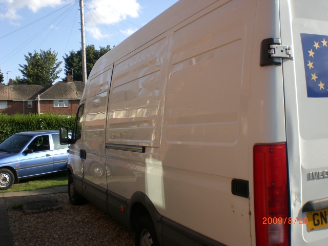 Biały samochód dostawczy Iveco Daily z 2004 roku, widok z tyłu, z naklejką z gwiazdami UE. Na tablicy rejestracyjnej widoczna data 2009/8/20. W tle niebieski samochód osobowy i domy.