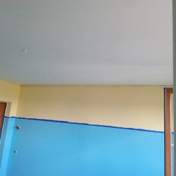 Dankar-Bud - Świeżo pomalowane wnętrze pokoju z dwukolorową ścianą: błękit na dole, kremowy u góry, oddzielone taśmą malarską. Biały sufit i widoczny fragment drzwi.