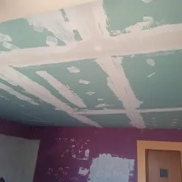 Montaż płyt kartonowo-gipsowych na suficie, widoczne łączenia wzmocnione taśmą i pokryte szpachlą, ściany częściowo pomalowane na fioletowo, widoczny fragment ościeżnicy drzwiowej.