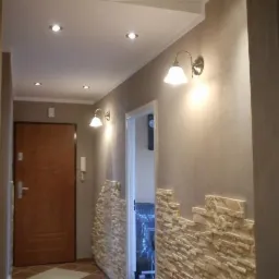 Korytarz po remoncie z dekoracyjną ścianą z kamienia, oświetleniem punktowym i kinkietami, brązową podłogą w karo i drzwiami wejściowymi.